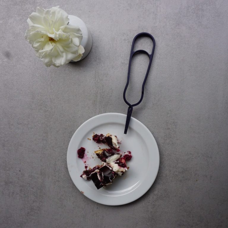 Ein Stück Kuchen auf einem weißen Teller, daneben eine Blume in einer Vase und das Lomega Besteck.